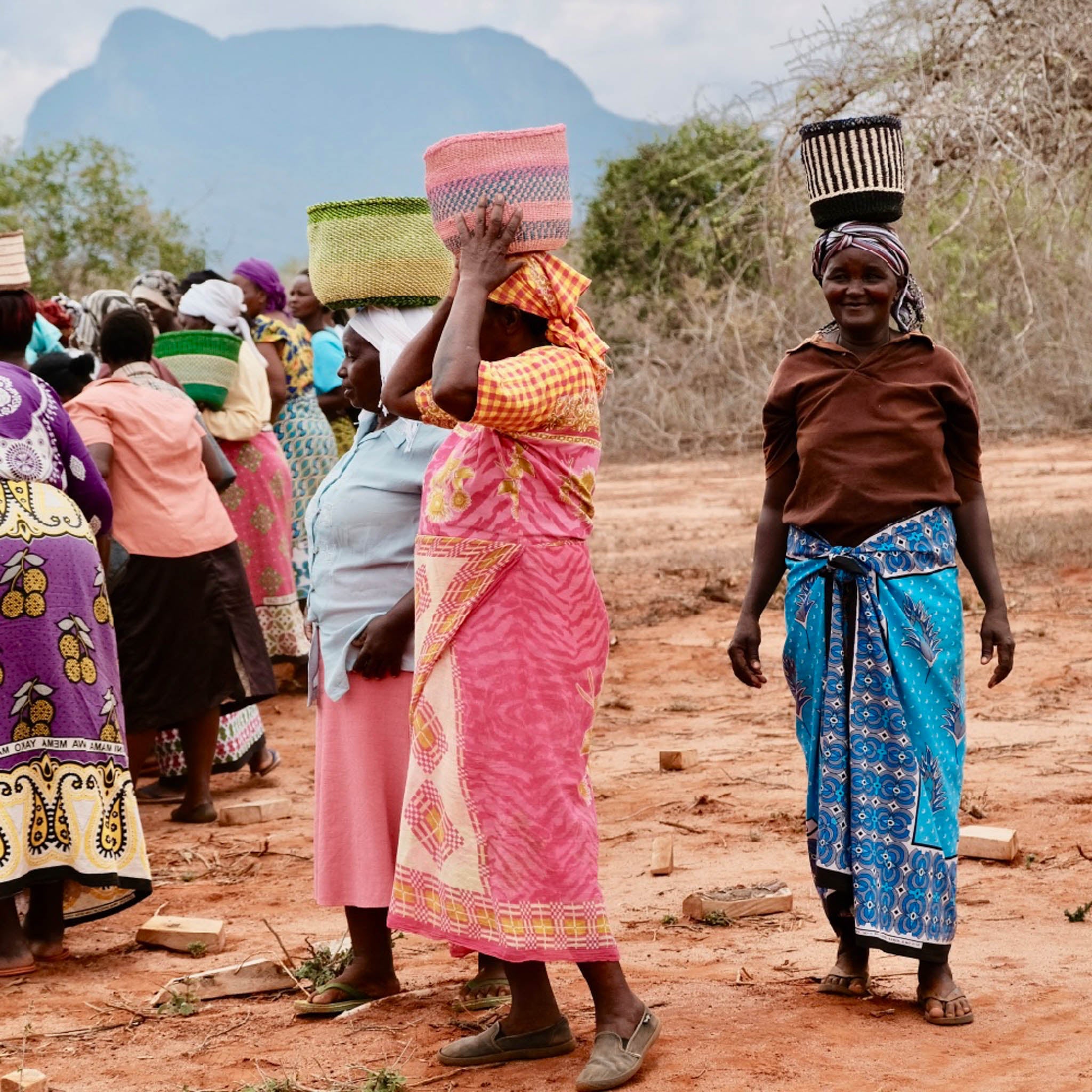 Women carrying baskets on their heads in a rural setting with mountains in the background.